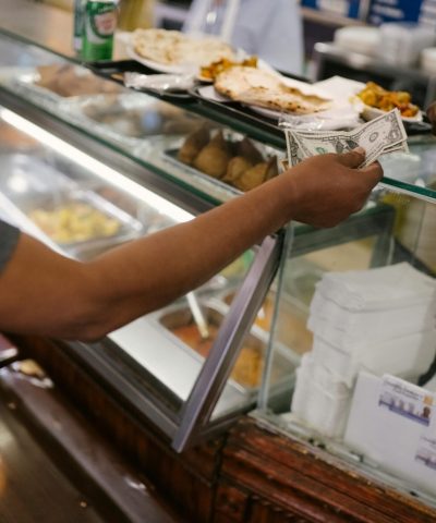 Crop faceless person standing near checkout buying food and passes money for dinner in cafe